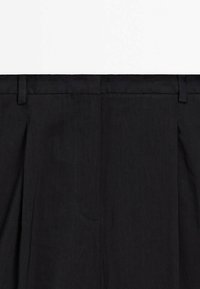 Pantaloni in tessuto nero con vita dotata di passanti per cintura, piega frontale singola e tasca laterale. Texture liscia con dettagli di cucitura minimi.
