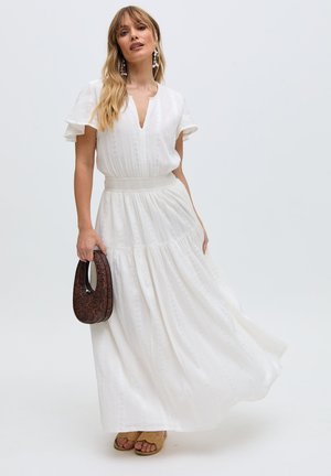 Frau in weißem knöchellangem Kleid mit flatternden Ärmeln, die eine braune, strukturierte Handtasche hält, trägt beige Sandalen und Tropfenohrringe.
