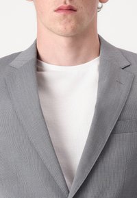 Blazer gris texturé avec col cranté, poche simple sur le côté gauche, porté sur un t-shirt blanc à col rond.