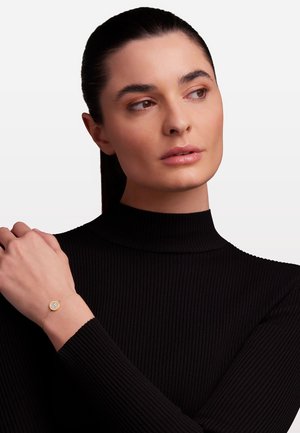 Goldfarbener Armreif mit einem runden Kristall-Mittelstück, zartem Kettchen und glatter Oberfläche, getragen am Handgelenk über einem strukturierten schwarzen Oberteil.