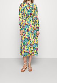 Blommig midiklänning med flerfärgade mönster i gult, blått och rosa. Långa ärmar och midjan är åtstramad, tillsammans med sandaler med remmar.