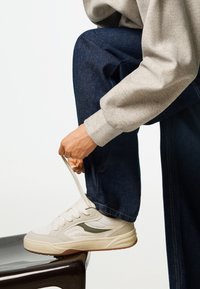 Fehér sportcipők struktúrált anyagból, szürke oldalsó akcentussal, vastag talppal és kerek orral, sötét farmernadrággal és szürke pulóverrel párosítva.