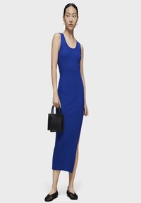 Vestido midi azul de canalé con escote redondo, abertura lateral y silueta ajustada. Combinado con un pequeño bolso negro y zapatos planos.
