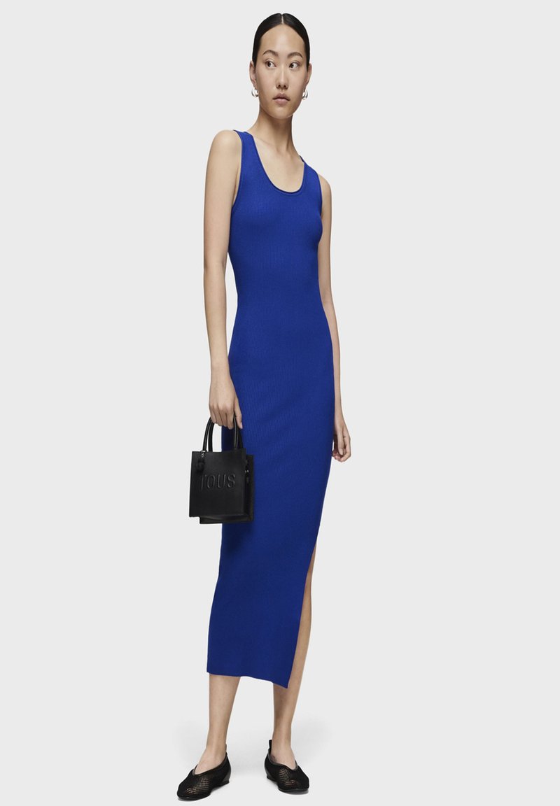 Vestido midi azul de canalé con escote redondo, abertura lateral y silueta ajustada. Combinado con un pequeño bolso negro y zapatos planos.
