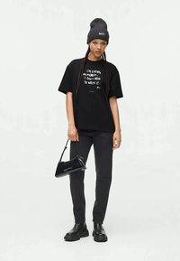 T-shirt gráfico preto com texto branco, calças skinnies pretas, botas de couro pretas e uma pequena bolsa preta. Um gorro completa o visual.