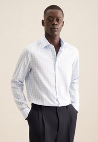 Camicia a quadri di colore blu chiaro realizzata in un tessuto intrecciato, con colletto, chiusura a bottone e maniche lunghe, abbinata a pantaloni scuri.