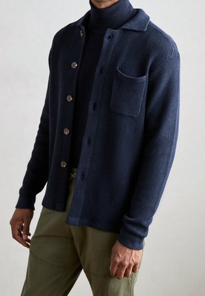 Homme portant un cardigan bleu marine texturé boutonné sur un col roulé foncé et un pantalon vert olive sur un fond uni.