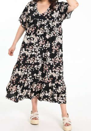 Femme portant une robe midi noire à motif floral blanc et rouge, avec des sandales à plateforme beiges, debout avec un bras levé.