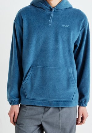 Person trägt eine blaue Fleece-Hoodie von Levi's mit einer Kängurutasche vorne und graue Hose, steht vor einem schlichten Hintergrund.