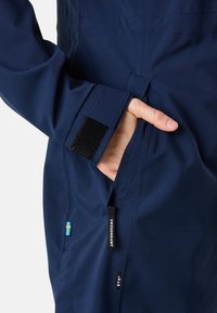 Marineblauwe waterdichte jas met klittenband manchetten, zichtbare zak en merklabel. Heeft een detail van het Zweedse vlag ontwerp op de stof.