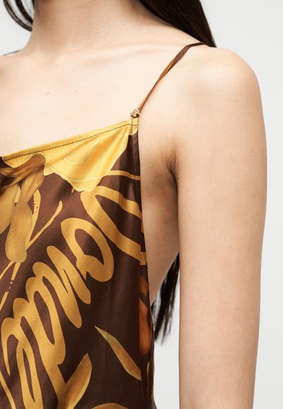 Κλείσιμο πλάνου μιας γυναίκας που φοράει φόρεμα με ραφή σπαγγέτι και καφέ και κίτρινο μοτίβο, που δείχνει τον ώμο και το πάνω μέρος του βραχίονα.