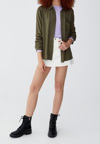 Chemise boutonnée vert olive, haut à manches longues violet clair, jupe en denim blanc avec ourlet effiloché, et bottines noires à lacets.