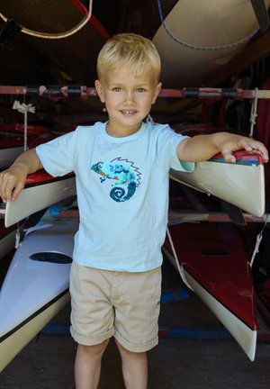 Hellblaues T-Shirt mit einem Pailletten-Chamäleon-Design, kombiniert mit beigefarbenen Shorts. Im Hintergrund sind rote Kajaks und weiße Kanus zu sehen.