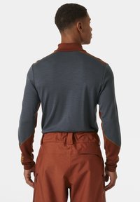 Langarm, grijze top met oranje accenten en een hoge kraag. Het materiaal lijkt lichtgewicht met een gestructureerde afwerking. Gecombineerd met oranje broeken.