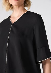 Svart bluse med V-hals, korte ermer med rynkedesign og hvit kant langs halslinningen og ermekantene. Glatt stoffstruktur.