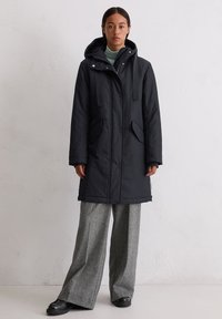 Czarny, ocieplany płaszcz z kapturem, z bocznymi kieszeniami i regulowanymi paskami, w zestawieniu z szarymi spodniami w szerokim kroju i czarnymi butami.