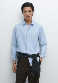 Lichtblauwe button-up shirt met een relaxte pasvorm en lange mouwen, gecombineerd met donkerbruine broek en een donkergrijze trui om de taille geknoopt.