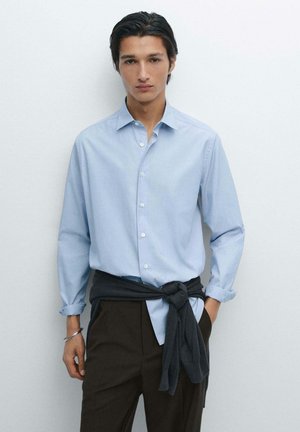 Lichtblauwe button-up shirt met een relaxte pasvorm en lange mouwen, gecombineerd met donkerbruine broek en een donkergrijze trui om de taille geknoopt.