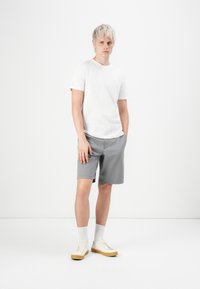 Jeune homme aux cheveux blonds portant un t-shirt blanc, un short gris, des chaussettes blanches et des baskets blanches, se tenant contre un fond blanc uni.