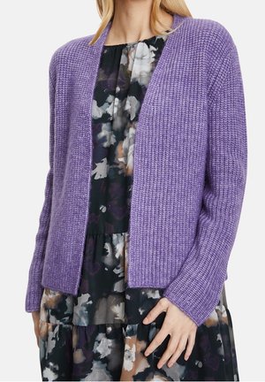 Femme portant un cardigan en tricot violet clair par-dessus une robe noire à fleurs avec des manches longues et une coupe ample.