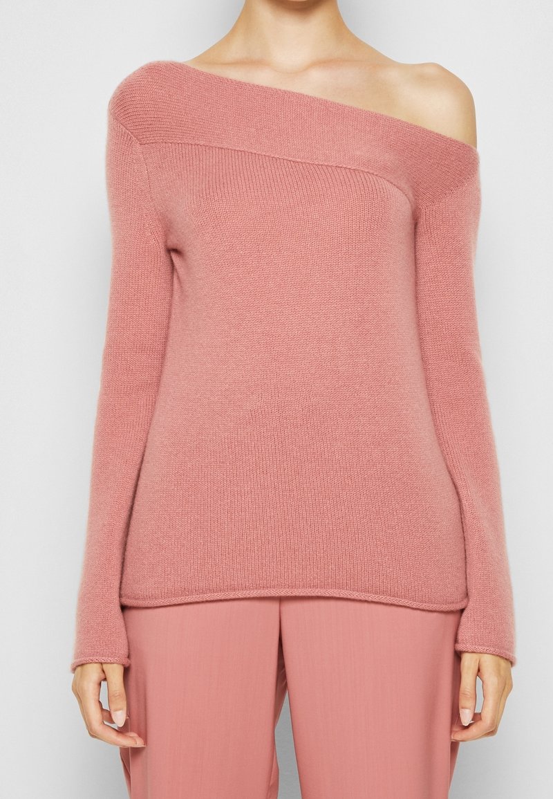 Pull en tricot rose à épaules dénudées avec des manches longues, présentant une texture douce et un col côtelé. Associé à un pantalon rose assorti.
