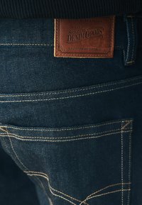 Jeans de mezclilla oscura con un parche de cuero marrón que dice "Next Signature Denim Goods", con costuras amarillas contrastantes en las costuras de los bolsillos.