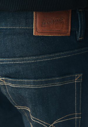 Donkerdenim jeans met een bruin leren label met de tekst "Next Signature Denim Goods", voorzien van contrasterende gele stiksels op de zaknaden.