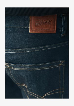Sötét farmernadrág, amelyen egy barna bőrpazító található, a "Next Signature Denim Goods" felirattal, ellentétes sárga varrással a zsebek varrásain.