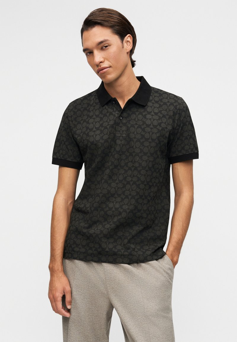 Man wearing a black patterned polo shirt with short sleeves and light gray pants, standing with one hand in pocket.