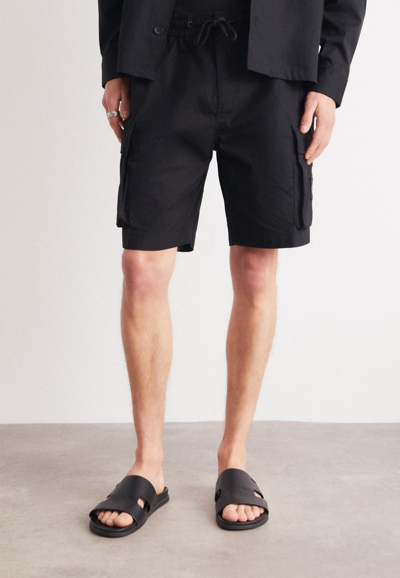 Personne portant un short cargo noir avec des poches latérales, une chemise noire, des bagues aux doigts et des sandales noires ouvertes à l’avant, debout sur un sol gris.
