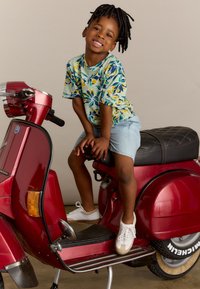 Trottinette rouge avec une finition mate, siège noir avec couture en losanges. Un enfant en chemise colorée à fleurs et en shorts bleu clair est assis dessus.