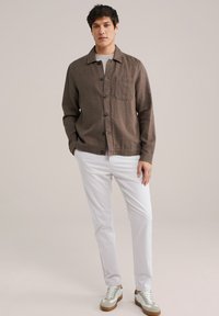 Brown linen button-up shirt with a chest pocket, paired with white trousers and grey sneakers. Simple design with a relaxed fit.
