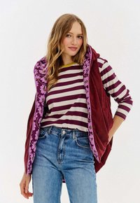 Młoda kobieta z luźnymi falującymi włosami, nosząca fioletową pasekowaną bluzkę z długim rękawem, bordową kamizelkę z kwiatowym podszyciem i niebieskie dżinsy na białym tle.