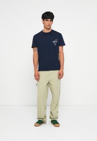 T-shirt en coton bleu marine à manches courtes avec un logo sur le devant de la poitrine ; associé à un pantalon ample vert clair et des baskets vertes.