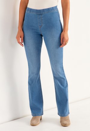 Lichtblauwe flared jeans met een gladde textuur en elastische tailleband, voorzien van gestikte naden en een ontspannen pasvorm. Gecombineerd met beige enkelboots.