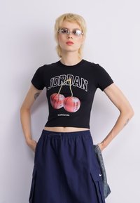 Black cropped t-shirt featuring a graphic design with basketballs, and "JORDAN" in bold white letters. Model styled with navy cargo shorts.