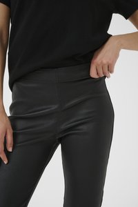 Černé kalhoty z faux kůže s vysokým pasem, hladkou texturou, elegantním designem a přiléhavým střihem, částečně zobrazené s rukou na pásku.