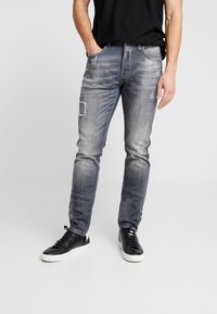 Jeans de mezclilla gris deslavado con acentos desgastados, con detalles de parches, diseño de corte slim y cinco bolsillos. Llevados con zapatillas negras.