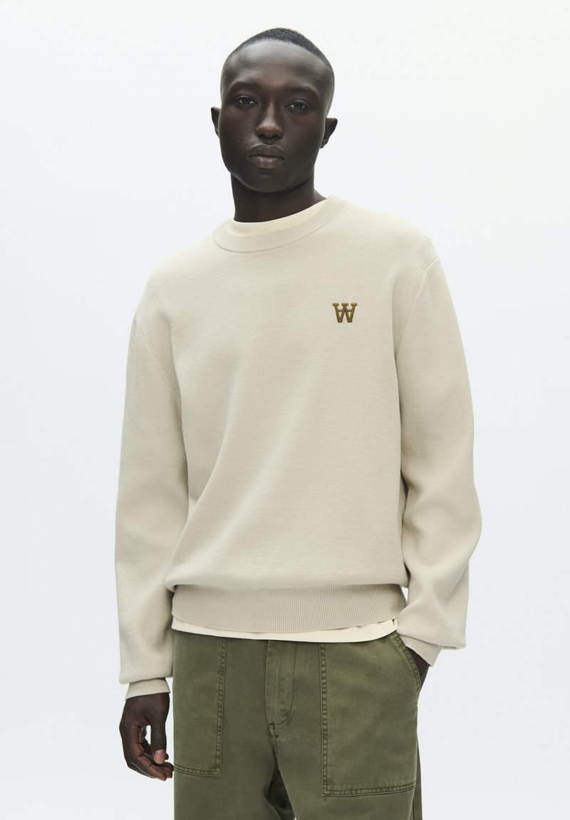 Sweat-shirt à col rond beige clair en tissu doux, avec poignets et ourlet côtelés, arborant un petit logo "W" marron sur la poitrine.