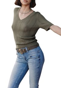 Pull à col V, à manches courtes, de couleur vert olive avec une texture côtelée, associé à un jean bleu clair et une ceinture brune avec une boucle dorée.