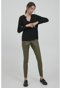 Svart blus med långa ärmar och v-ringning, kombinerad med olivgröna, figursydda byxor i ett glansigt material. Svarta loafers fulländar outfiten.