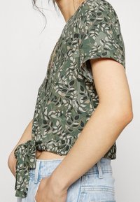 Top corto floreale verde con maniche corte, caratterizzato da un profondo scollo a V e un dettaglio con nodo in vita, realizzato in tessuto leggero.
