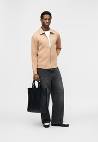 Άνδρας με μπεζ μπουφάν με φερμουάρ, λευκό πουκάμισο, άνετο μαύρο τζιν, μαύρα και λευκά παπούτσια κρατώντας μια μαύρη δερμάτινη τσάντα tote στέκεται σε λευκό φόντο.