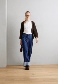Bruine cropped cardigan, witte blouse met knopen, hooggetailleerde donkerblauwe jeans met lichtdenim omslag, zwarte platformlaarzen, achtergrond is een gladde grijze muur.