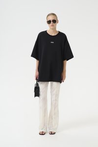 Svart oversized t-skjorte med logo på brystet, kombinert med hvite mønstrede vide bukser. Tilbehør inkluderer svarte solbriller og en liten håndveske.