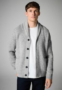 Šedý textúrovaný cardigan s šálov�ým golierom, šiestimi čiernymi gombíkmi a prednými vreckami, nosený cez biele tričko a spárovaný s tmavými džínsami.