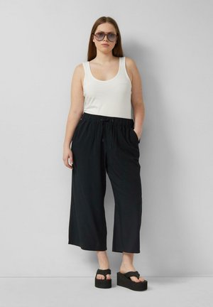 Donna con capelli lunghi con occhiali da sole con lenti colorate, top bianco senza maniche, pantaloni neri larghi e sandali neri con plateau, in piedi contro un muro grigio.