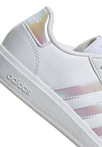 White sneakers with a textured rubber sole, smooth synthetic upper, and iridescent accents on the heel and three lateral stripes.