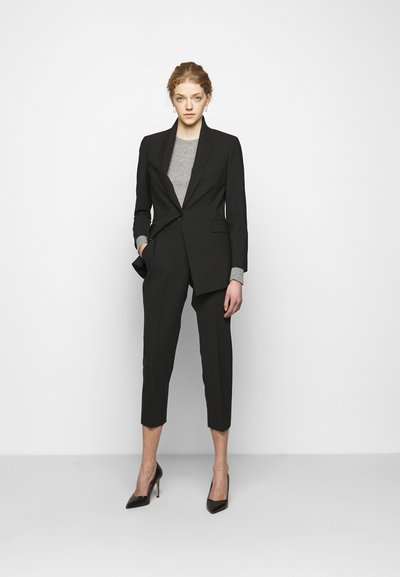 Fato preto sob medida com um corte ajustado, apresentando um blazer de um botão, calças curtas e um suéter cinza por baixo. Sapatos de salto fino pretos completam o visual.