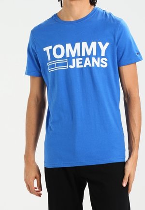 Blauw katoenen T-shirt met een gedurfde witte "TOMMY JEANS"-logo op de voorkant. Ronde hals, korte mouwen en standaard pasvorm. Zwarte broek zichtbaar.